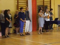 Zdjęcie przedstawia panią Dyrektor przemawiającą podczas uroczystości rozpoczęcia  roku szkolnego. Za nią, na tle falgi Polski, stoi grupa nauczycieli.